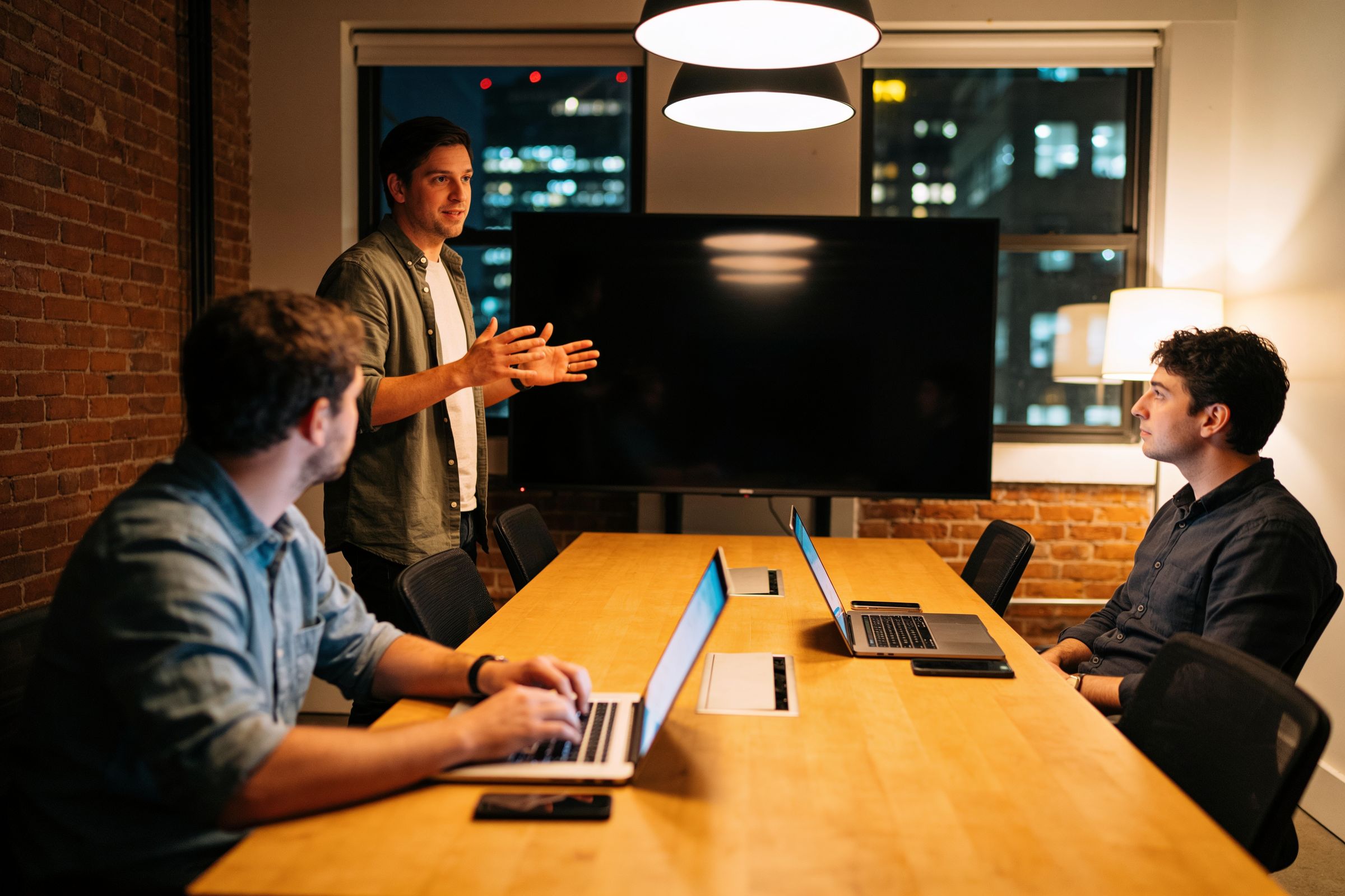 Three people in an agency meeting room at night, one presenting